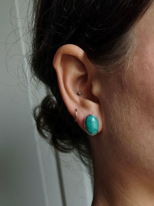 Close-up of a person's ear adorned with two earrings: a small sterling silver hoop in the helix and an Oval Turquoise Stud by Kathrin Jona in the lobe.