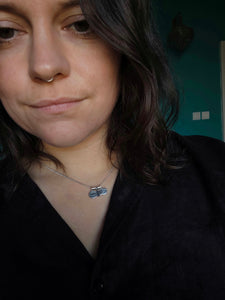 A person with dark hair and a nose ring stands in front of a teal wall, wearing a black shirt. Around their neck is a necklace featuring the 'YIN YANG' silver charm by Kathrin Jona, adding an intriguing element to their look.