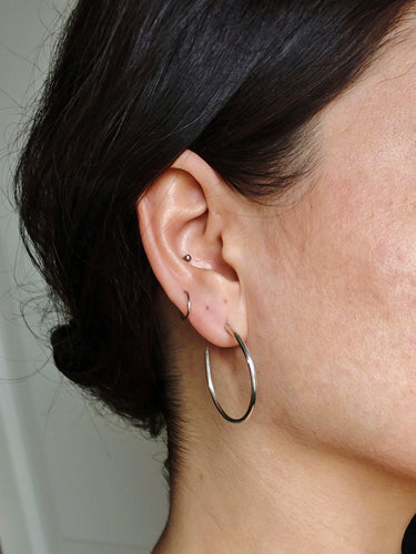 A close-up of a person's ear adorned with Kathrin Jona's Silver Round Hoop Hinge Earrings and a small stud in the cartilage.