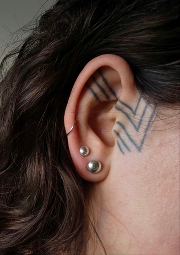 The close-up showcases a person's ear decorated with Kathrin Jona's Recycled Silver Ball Studs in various sizes, complemented by geometric tattoos stretching from the temple to the ear. The area is partially veiled by brown hair.