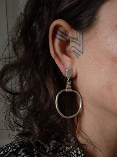 A person with curly hair flaunts a large Matte Organic Hoop earring by Kathrin Jona in recycled sterling silver, featuring an organic shape, alongside a geometric tattoo behind the ear and wearing a patterned shirt.