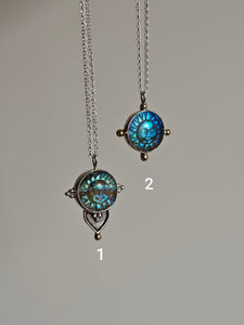 Explore two sun-themed necklaces on chains: The first features a teardrop pendant crafted from recycled silver with an ornate design, while the second, the Kathrin Jona Fair Labradorite Sun Face Necklace, showcases a round blue sun face adorned with minimal upcycled gold accents.