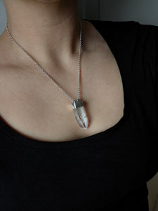 A person wears a black top, accessorized with the Australian Clear Quartz Necklace #2 by Kathrin Jona, an adjustable silver chain necklace crafted from recycled silver and featuring a clear quartz pendant.