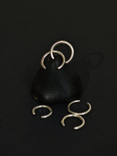 The Ear cuff by Kathrin Jona features a black stone pendant adorned with two attached silver rings, all crafted from recycled sterling silver and displayed upright. Accompanying it are three additional silver rings on the dark surface, creating an elegant and unique accessory ensemble reminiscent of ear cuffs.