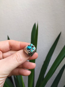 A hand holding a Kathrin Jona Turquoise Cluster Signet Ring in front of a plant.
