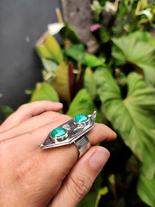 A hand holding a Green Shield Malachite Ring #3 by Kathrin Jona in front of plants.