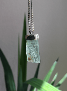 A piece of Aquamarine Necklace No.4 by Kathrin Jona on a silver chain hanging from a plant.